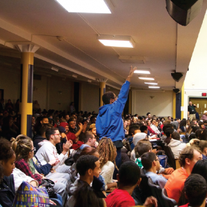 a crowded high school auditorium with one student standing up with hands raised